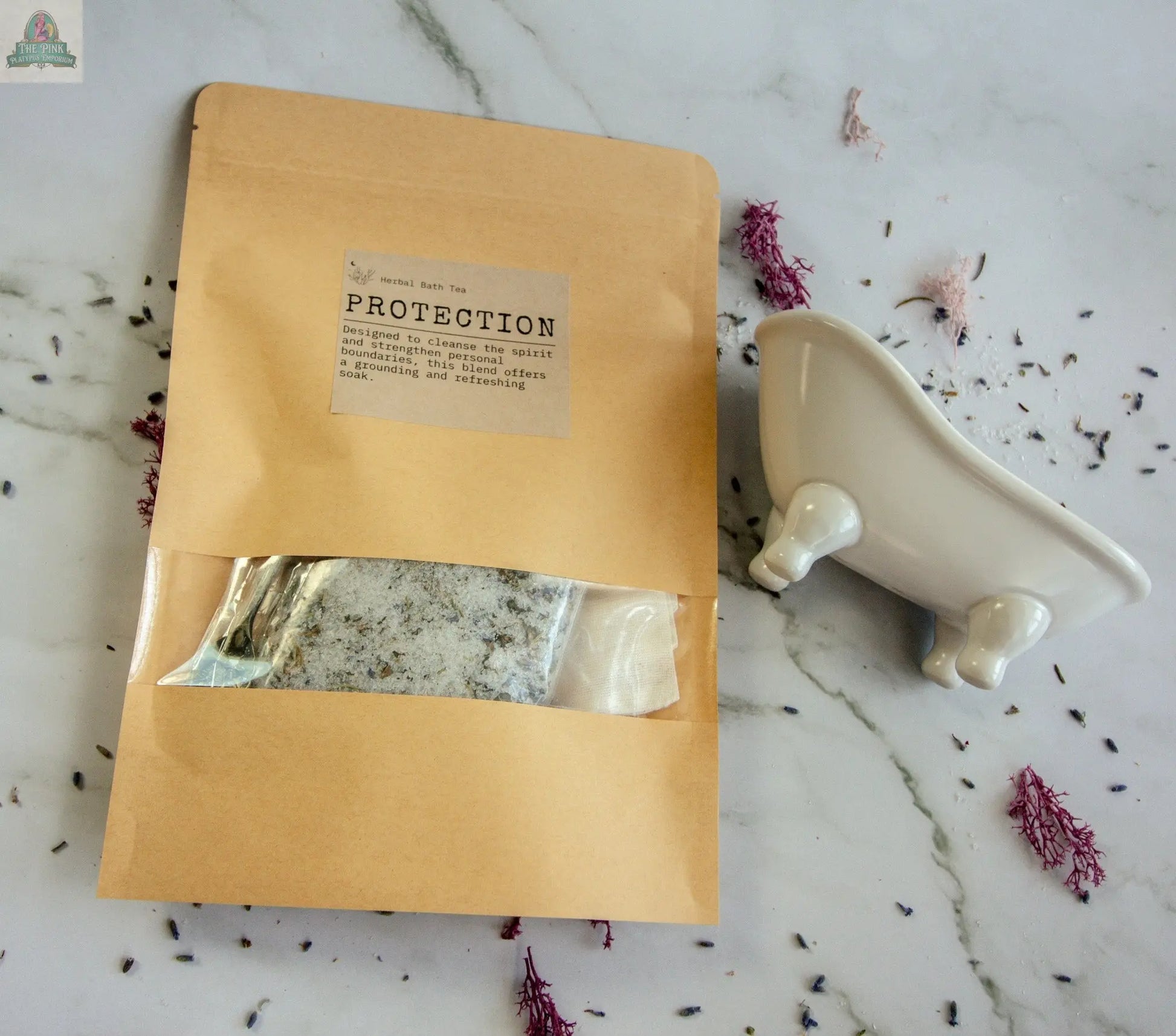 A brown paper pouch labeled Protection holds a sachet of herbal bath tea, shown on a marble surface with scattered lavender and a white clawfoot-tub-shaped soap dish beside it.