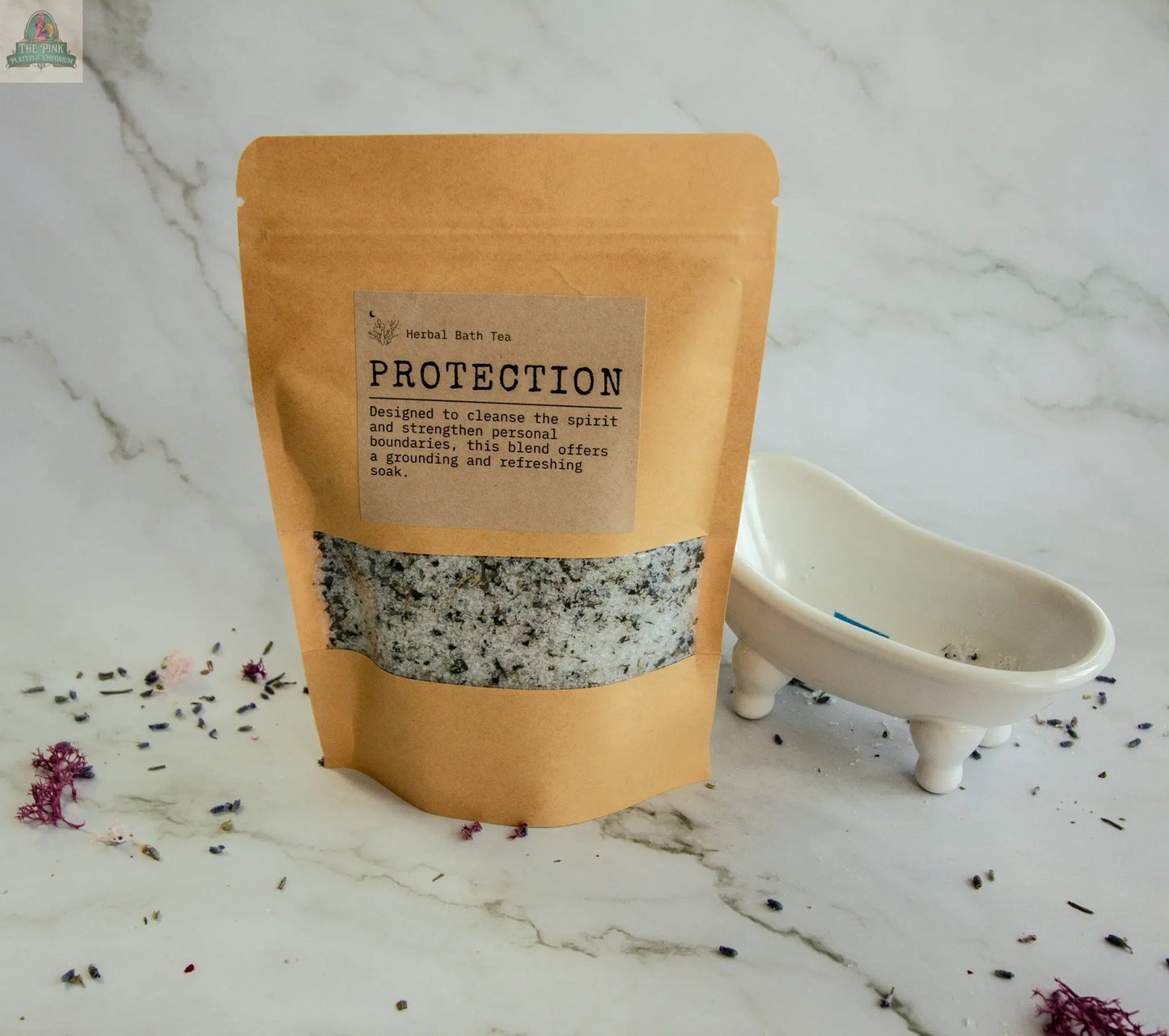 A kraft paper pouch labeled "Protection" herbal bath tea stands on white marble next to a small tub dish, with scattered dried herbs and petals—ideal for a protective bath soak with calming botanicals.