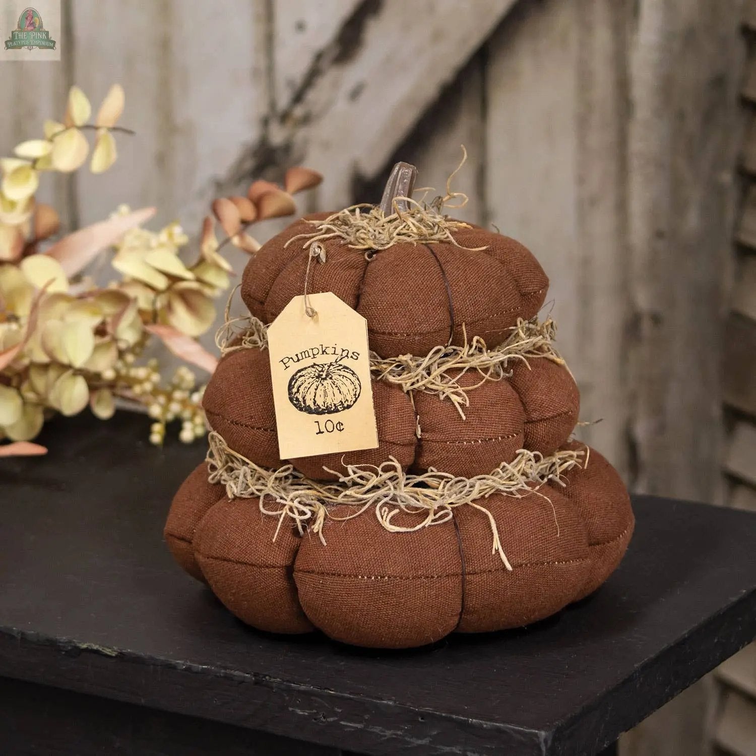 The "Pumpkins 10 Cents" Stuffed Pumpkin Stack features three brown plush pumpkins with straw accents and a tag, making it a charming rustic decoration for fall displays.