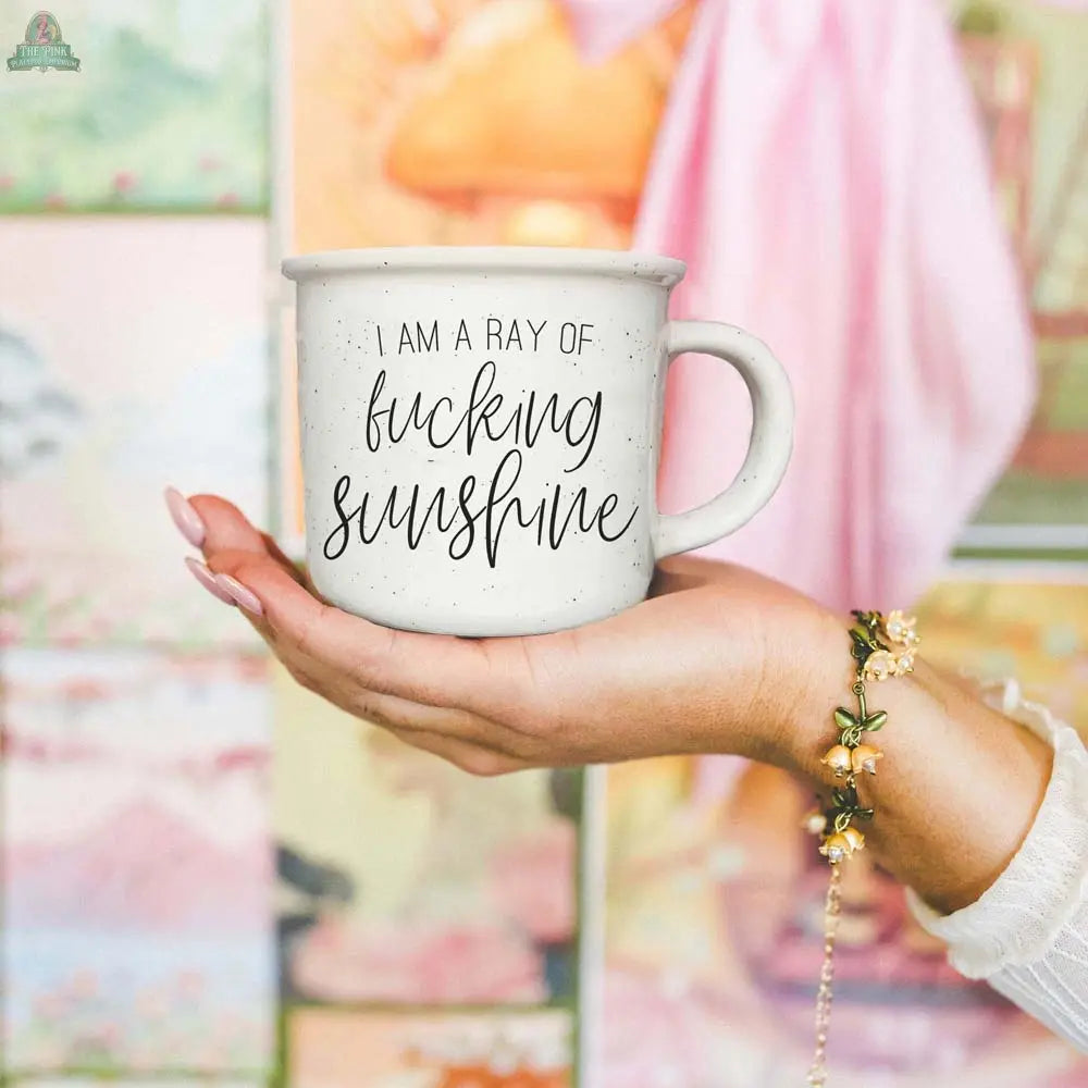A hand with a floral bracelet holds the Ray of F'n Sunshine 14.5oz mug, featuring "I am a ray of fucking sunshine" in black script, against a colorful blurred background.