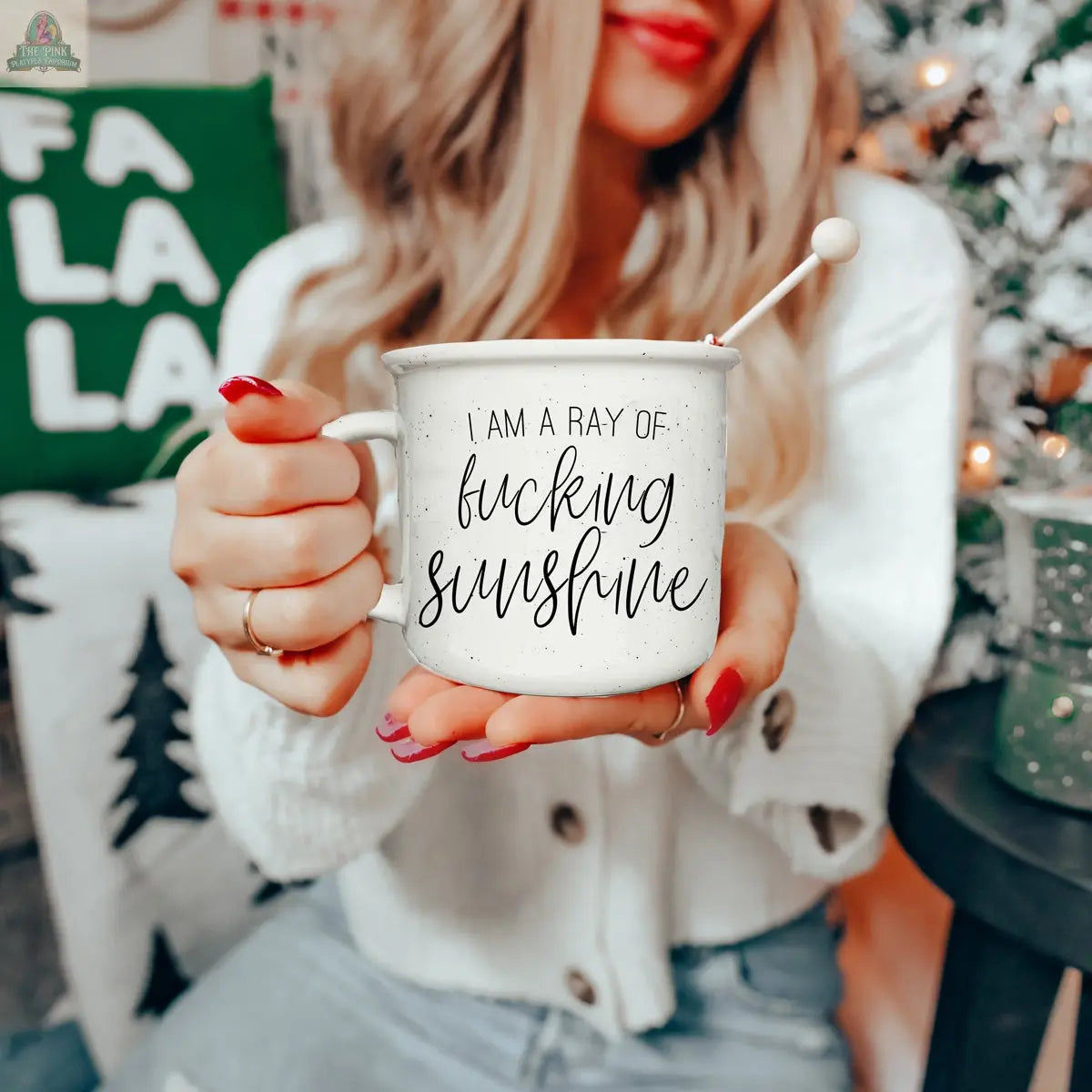 A woman with long blonde hair and red nails holds a Ray of F'n Sunshine 14.5oz mug. She wears a white sweater, and festive holiday decor is visible in the background.