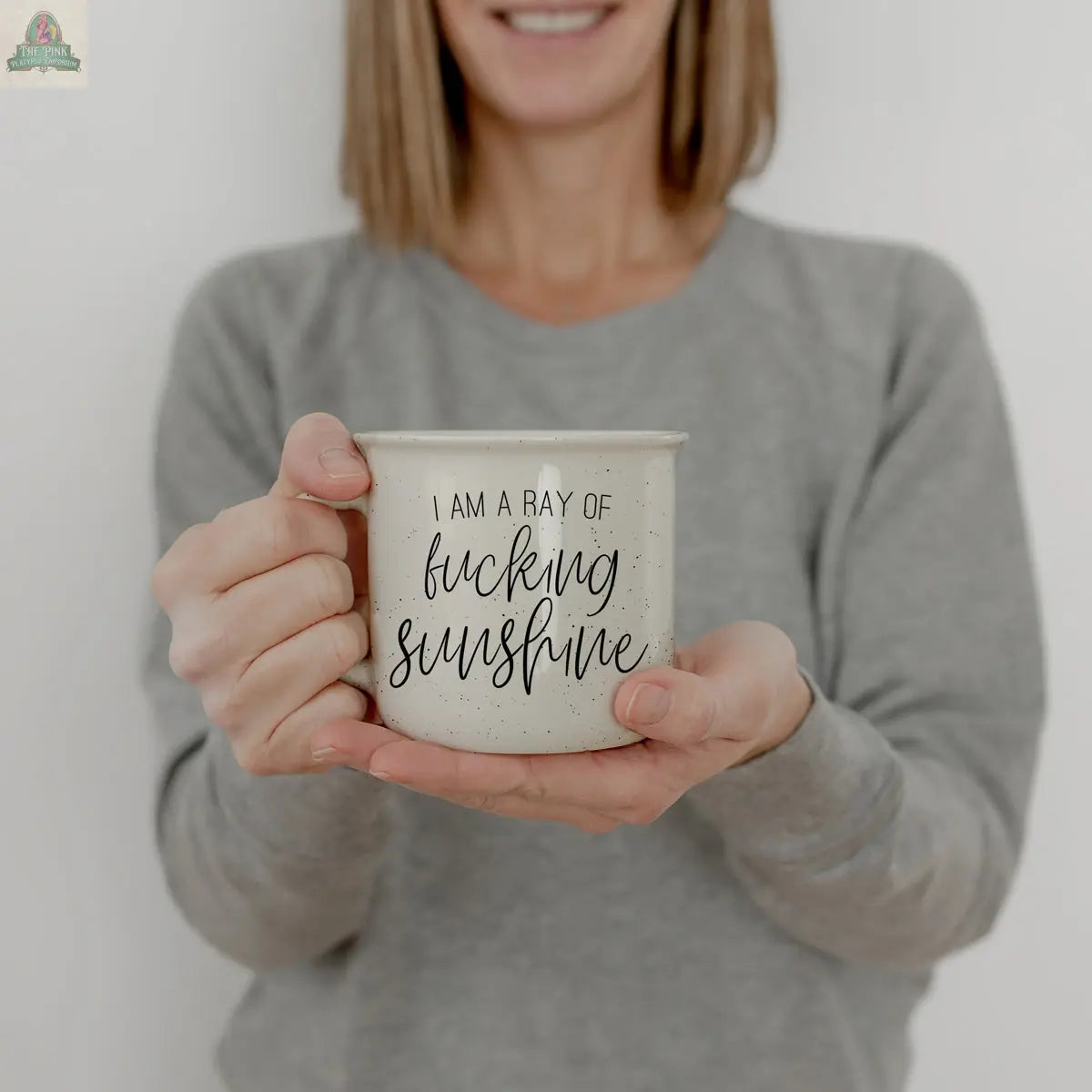 A woman in a gray sweater holds the Ray of F'n Sunshine 14.5oz ceramic mug with "I am a ray of fucking sunshine" printed on it. The background is plain and light-colored.