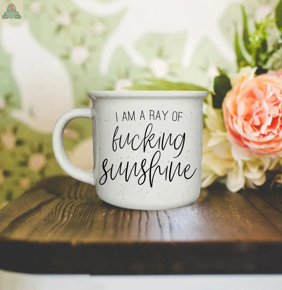 A white speckled Ray of F'n Sunshine 14.5oz ceramic mug sits on wood, featuring black script with pink flowers and a green patterned wall in the background.