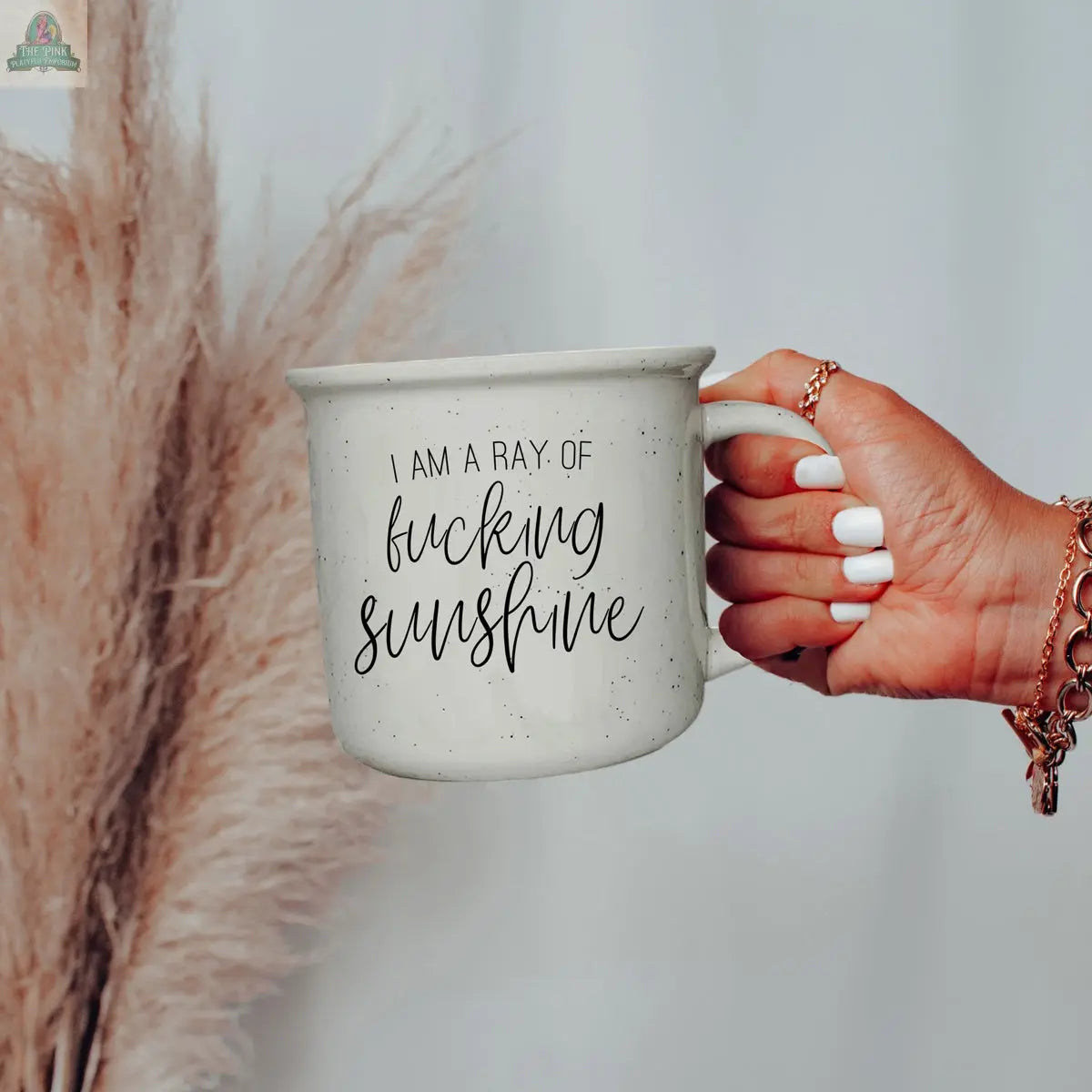 A hand with white nail polish and gold bracelets holds the Ray of F'n Sunshine 14.5oz mug, with pampas grass in the background.