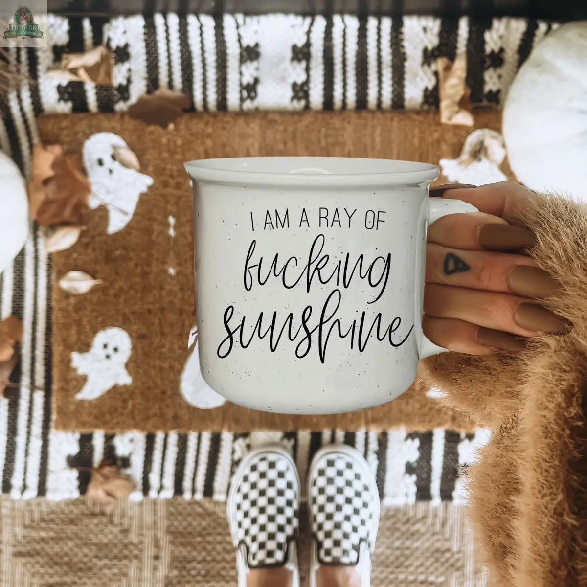 A person with manicured nails and a triangle tattoo holds a Ray of F'n Sunshine 14.5oz mug over a fall doormat with white ghost designs, wearing checkered slip-on shoes.