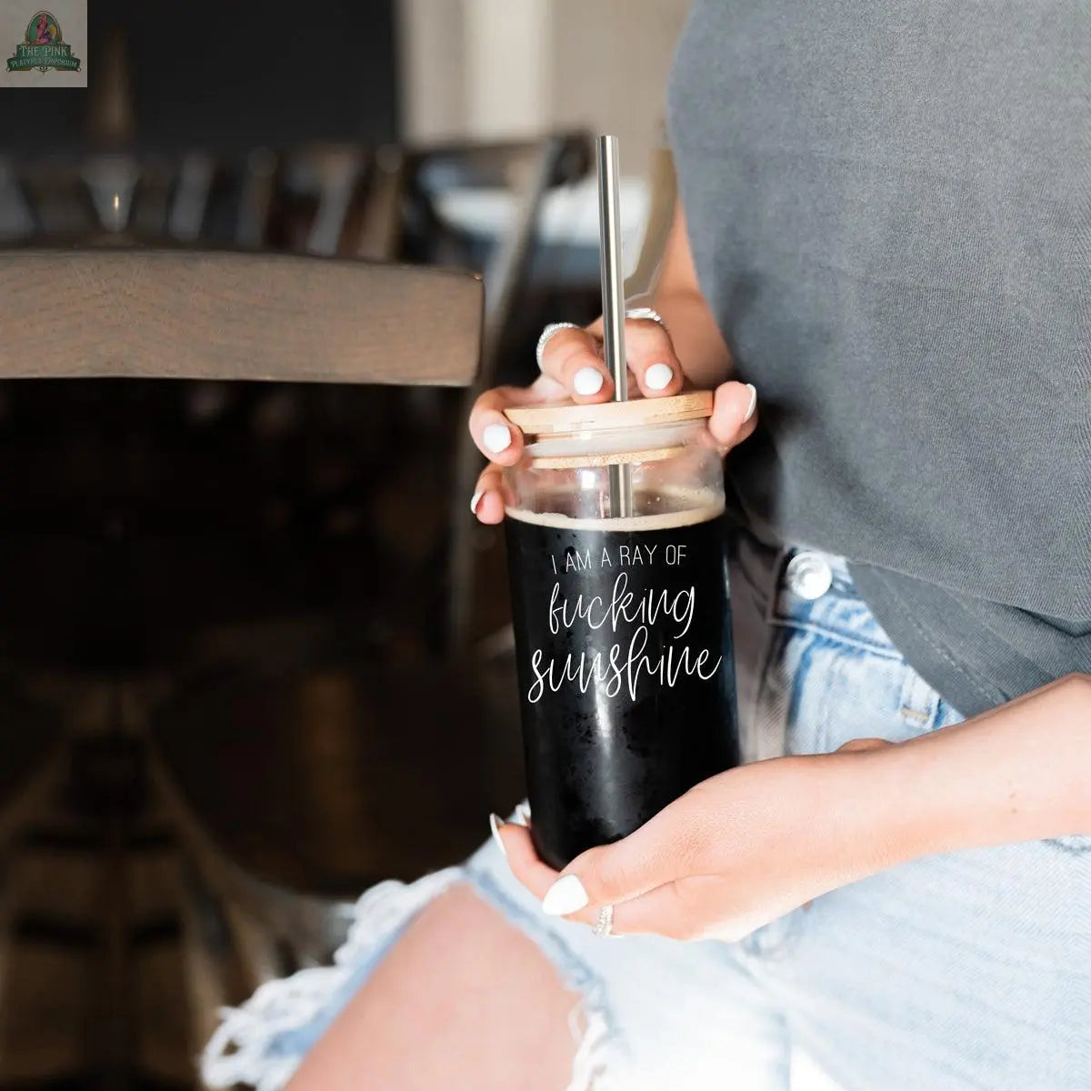 A person in a gray shirt and ripped denim shorts holds the Ray of Sunshine 17oz glass cup with a straw and dark iced coffee. The jar features "I am a ray of fucking sunshine" in white script.