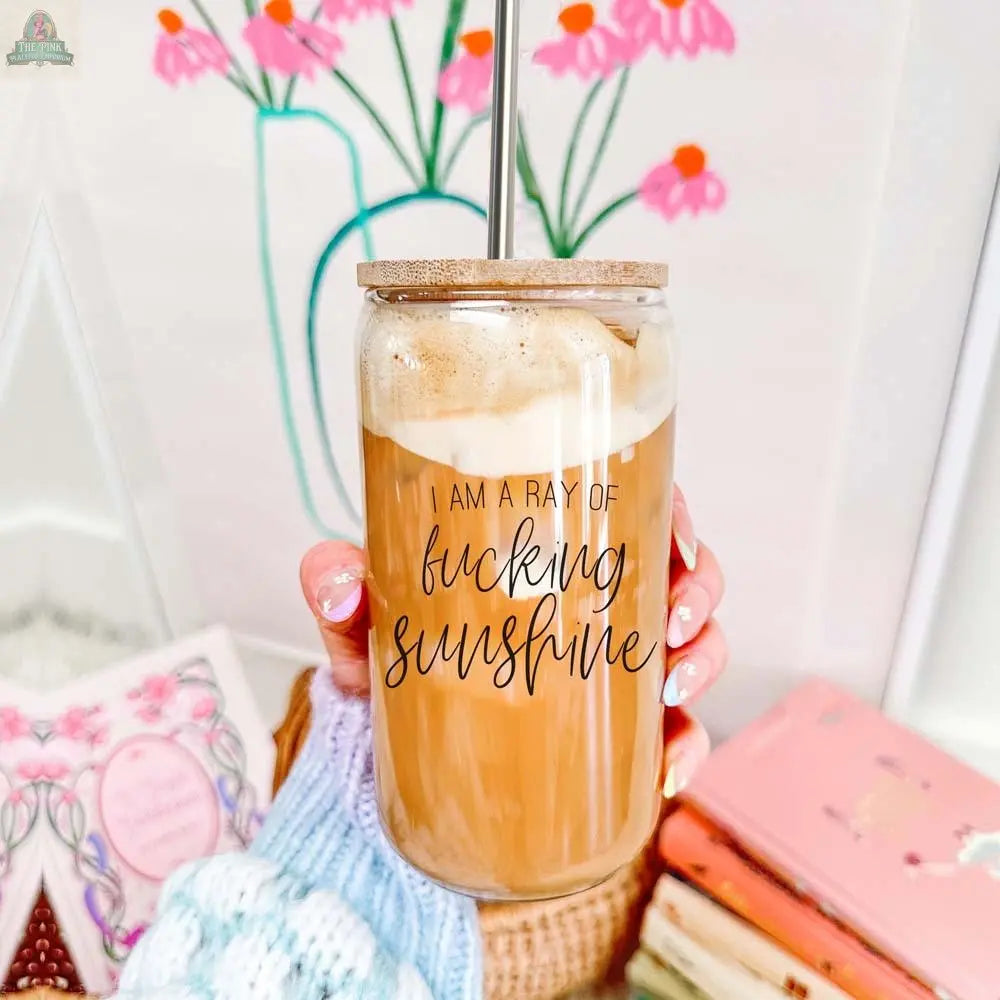 A hand holds a Ray of Sunshine 17oz glass cup filled with iced coffee, topped with a bamboo lid and straw. The glass features the phrase "I am a ray of fucking sunshine," with colorful books, a knit blanket, and a floral drawing in the background.
