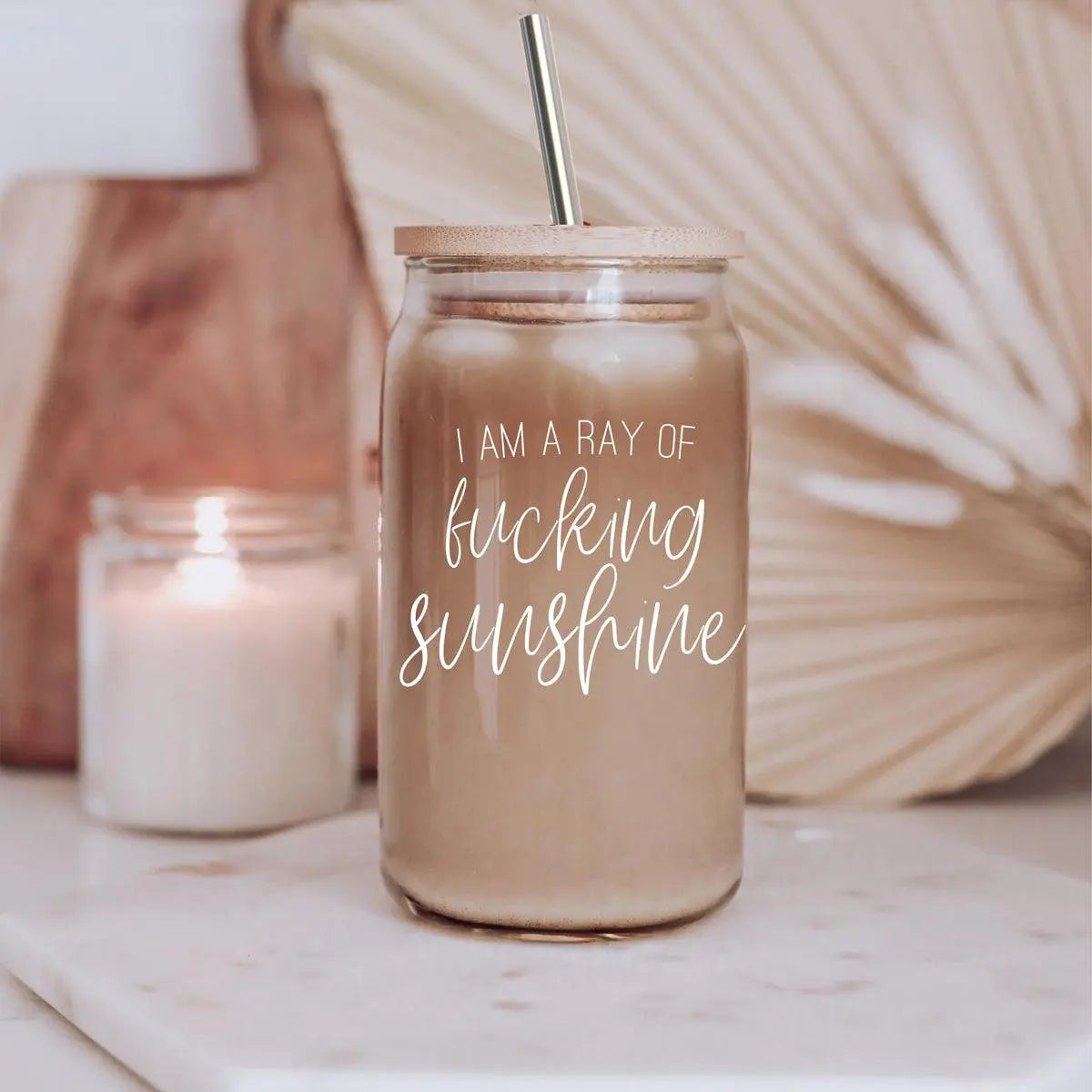 The Ray of Sunshine 17oz glass mason jar with bamboo lid and straw holds a creamy drink on a marble surface. “I am a ray of fucking sunshine” is printed in white script. A lit candle and neutral decor appear in the background.