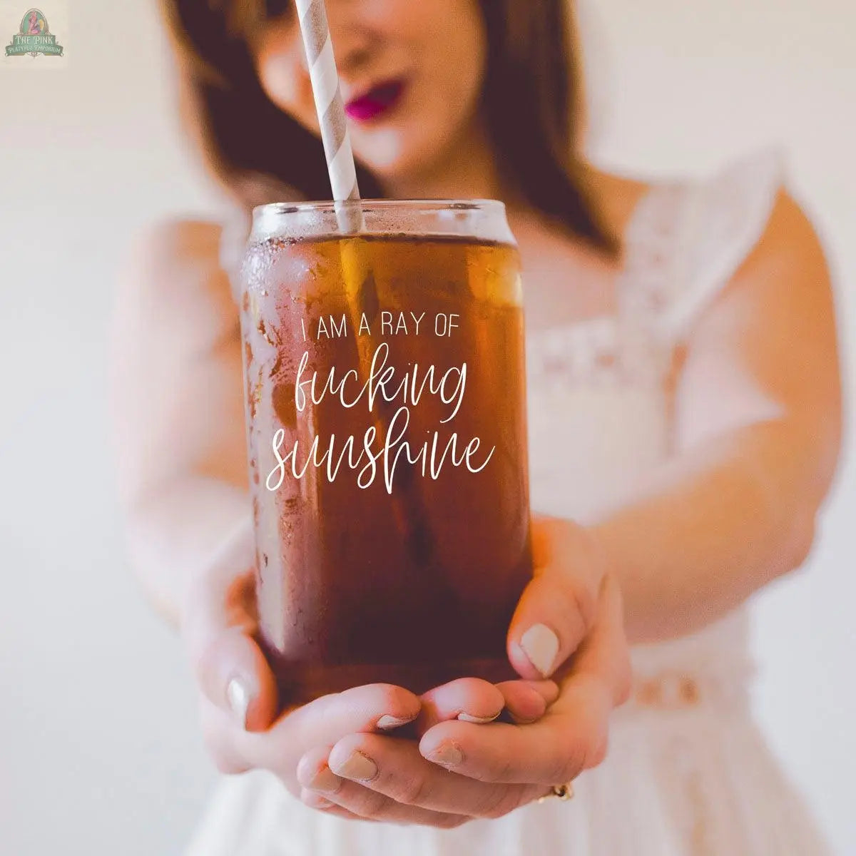 A woman in a white dress holds the 17oz Ray of Sunshine jar with a stainless steel straw, its bold "I am a ray of fucking sunshine" text standing out against a softly blurred background.