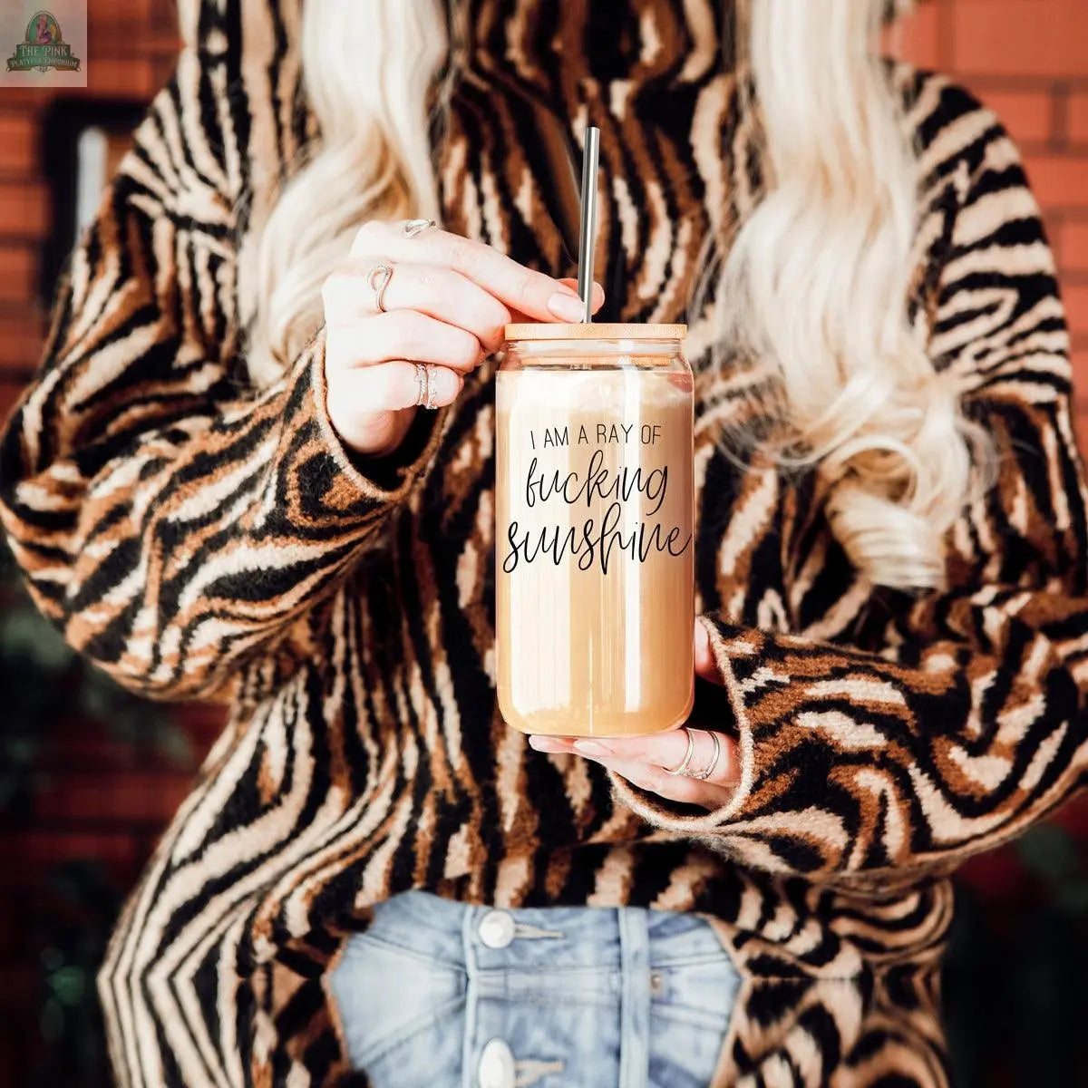 A person in a zebra-striped sweater holds a Ray of Sunshine 17oz glass cup with a straw and bamboo lid. The cup has the phrase “I am a ray of fucking sunshine.” Their long blond hair is visible, but not their face.