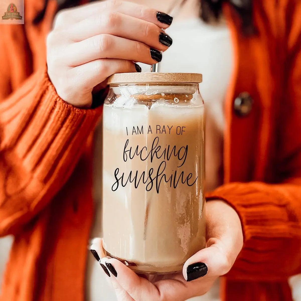 A person in an orange sweater holds the Ray of Sunshine 17oz glass jar with iced coffee. The jar has a bamboo lid, stainless steel straw, and black text saying, I am a ray of fucking sunshine.