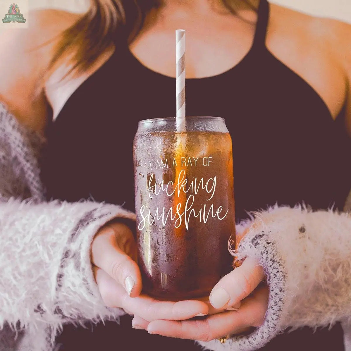 A woman in a black tank top and cozy sweater holds the Ray of Sunshine 17oz glass jar with a bamboo lid and striped straw, featuring the phrase “I am a ray of f*cking sunshine,” filled with what looks like iced tea or soda.