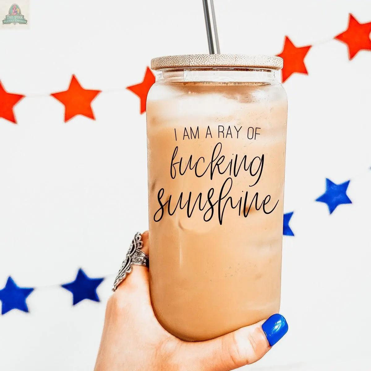A hand with blue nail polish holds an iced drink in the Ray of Sunshine 17oz glass cup with a stainless steel straw. Red and blue star garlands decorate the white background.