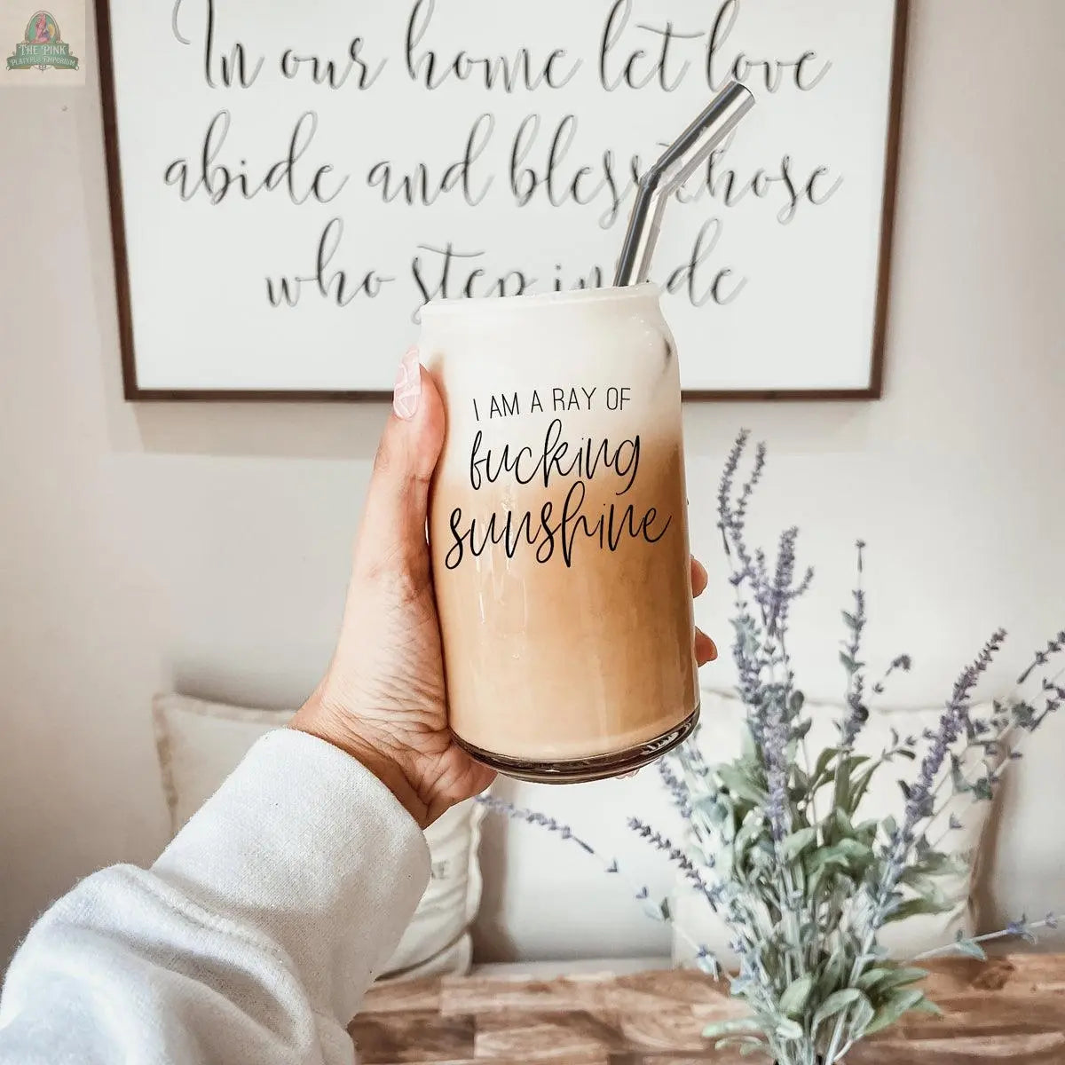 A hand holds the Ray of Sunshine 17oz glass cup filled with iced coffee, featuring a stainless steel straw. In the background, a decorative sign and lavender flowers are arranged on a wooden table.