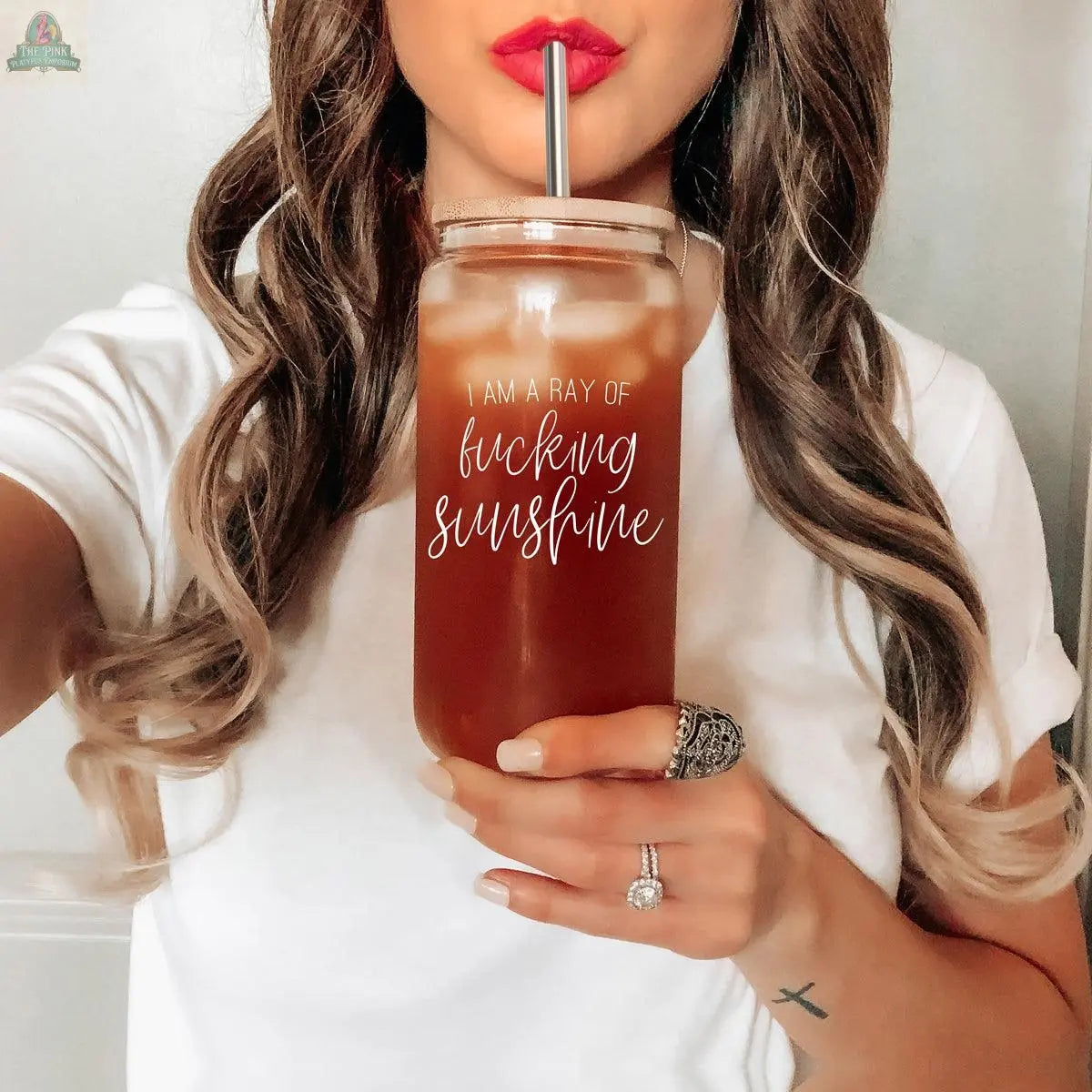 A woman with long wavy hair and bold lipstick holds the Ray of Sunshine 17oz glass, which reads "I am a ray of fucking sunshine." She wears a white shirt and rings, sipping through a stainless steel straw.