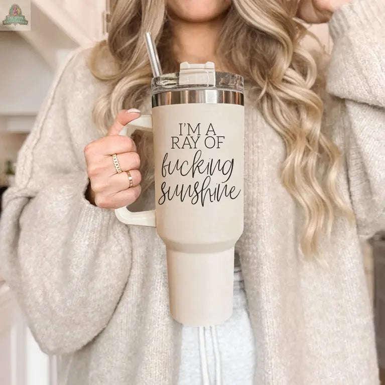 A woman with long, wavy blonde hair holds the Ray of Sunshine 40oz insulated mug, which has a handle and the phrase “IM A RAY OF f*cking SUNSHINE” in black script. She wears a beige cardigan over a gray top.