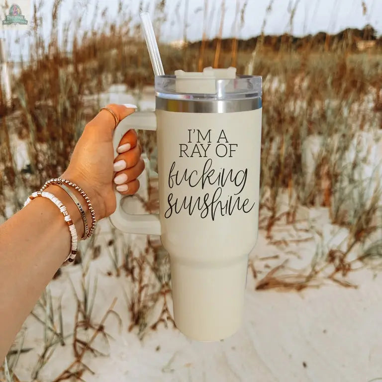 A hand holds the Ray of Sunshine 40oz insulated beige mug with lid and straw, reading "I'm a ray of fucking sunshine." Its beachside setting shows this mug is ideal for outdoor adventures.