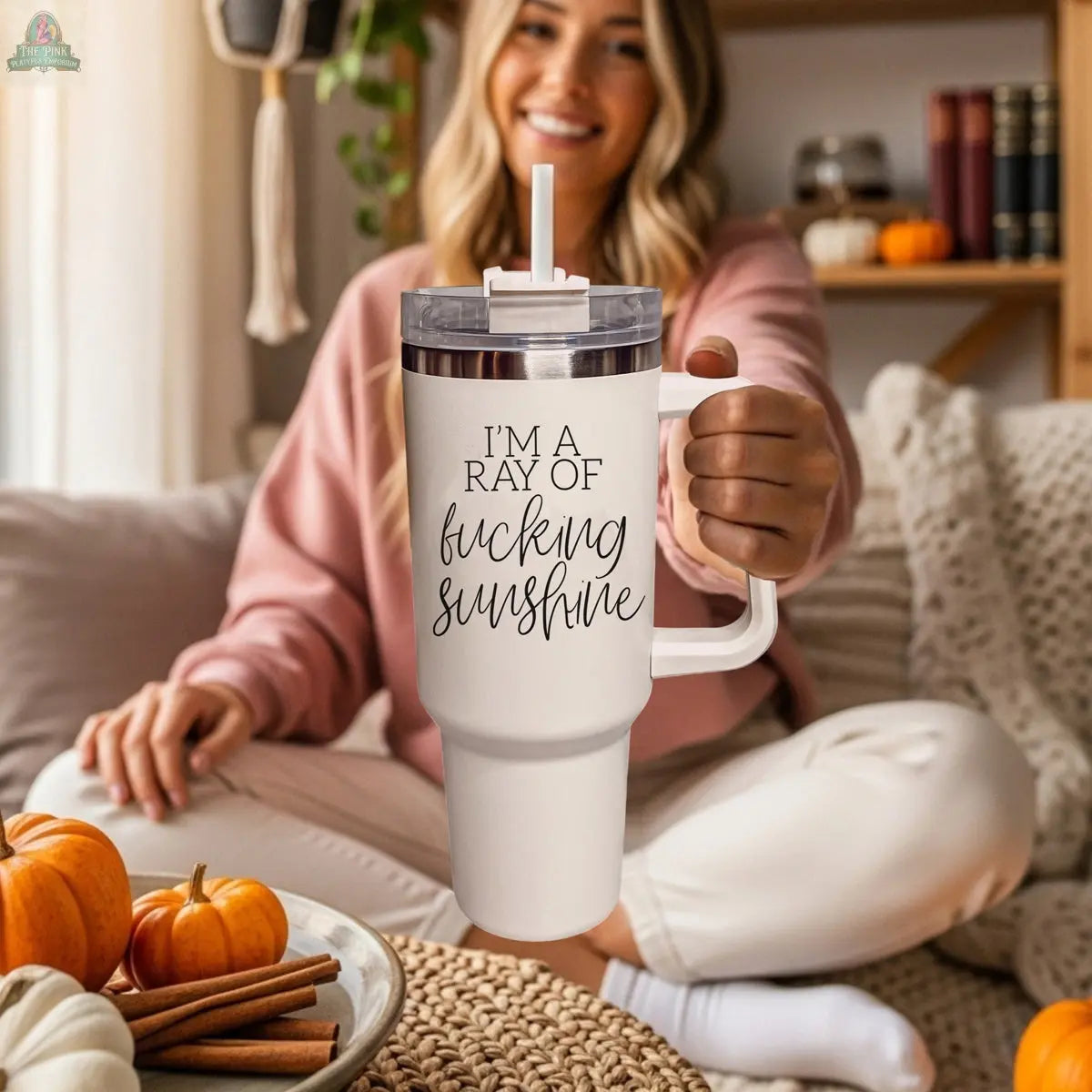 A smiling woman sits cross-legged on the floor, holding the Ray of Sunshine 40z insulated mug with “I'M A RAY OF f*cking SUNSHINE.” She’s in a cozy room decorated with pumpkins, books, and autumn-themed decor.