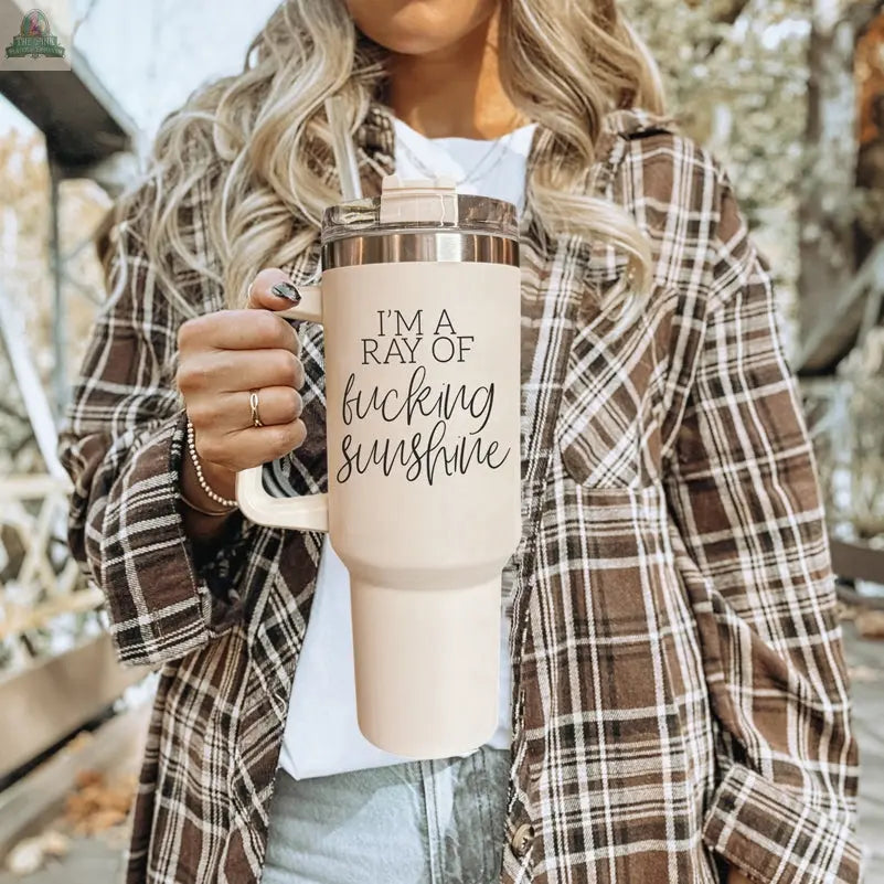 A woman in a plaid shirt holds the “Ray of Sunshine 40oz” insulated mug printed with “I’M A RAY OF fucking sunshine,” standing outdoors among trees and seating.