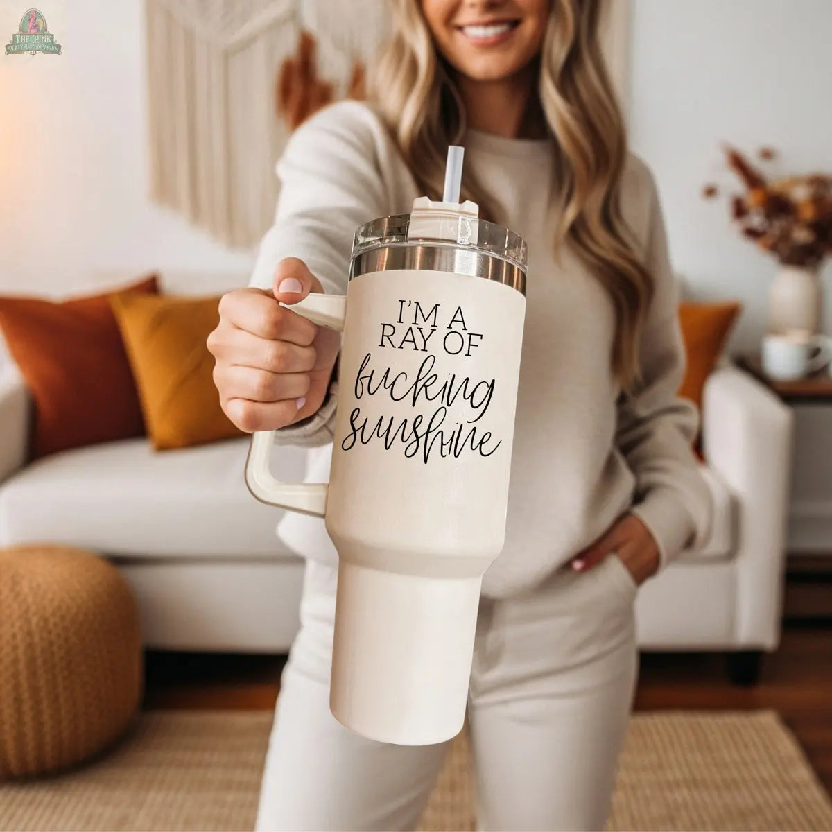 A smiling woman in neutral loungewear holds the Ray of Sunshine 40oz travel mug, featuring “I’m a ray of fucking sunshine,” while standing in a cozy, sunlit living room with warm accents.