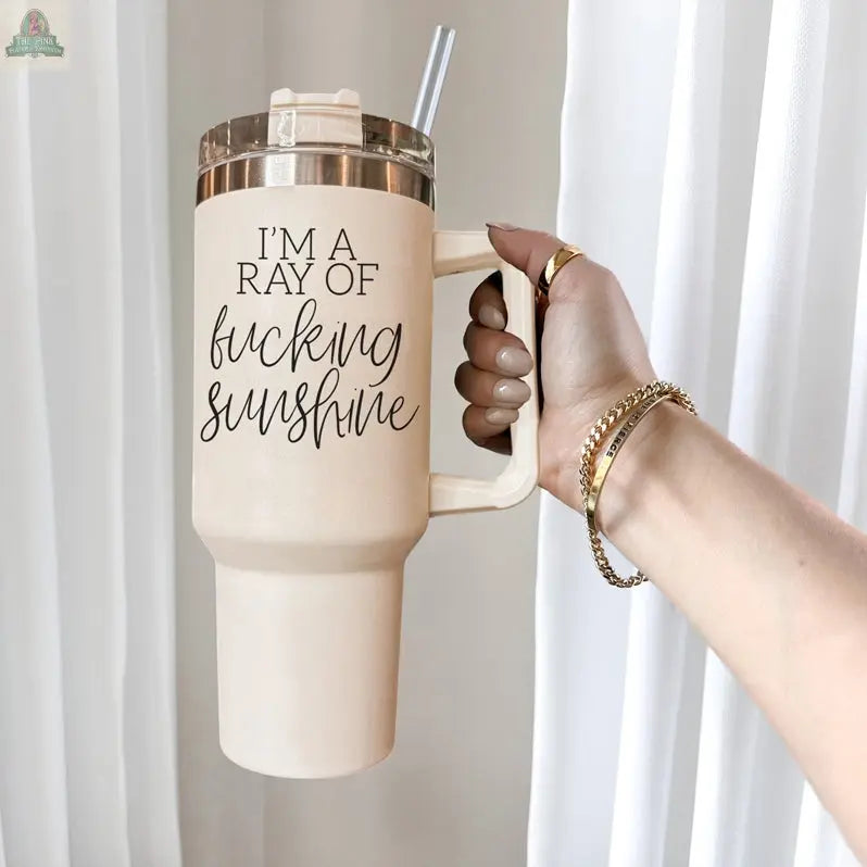 A hand with manicured nails and gold bracelets holds the Ray of Sunshine 40oz insulated mug with a lid and straw. The mug reads "IM A RAY OF fucking sunshine." White curtains hang in the background.