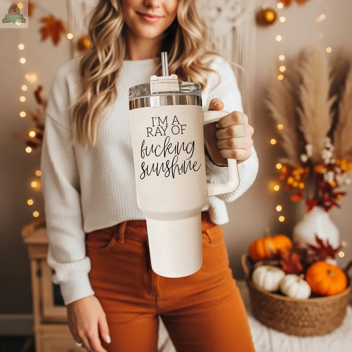 A woman in an autumn-themed room holds the Ray of Sunshine 40oz mug. Dressed in an off-white sweater and rust pants, she’s surrounded by pumpkins and cozy fall decor, showing off the mug’s bold “IM A RAY OF fucking SUNSHINE” print.