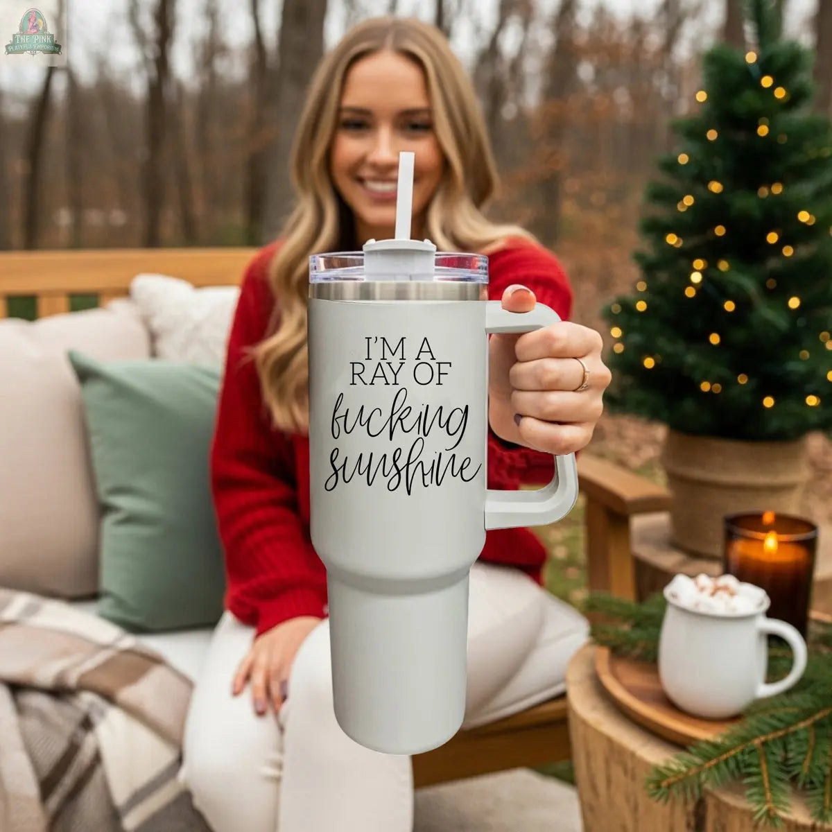 A smiling woman in a red sweater sits outdoors on a cozy patio, holding the "Ray of Sunshine 40z" insulated mug. Nearby are a small lit Christmas tree and a mug of hot chocolate.