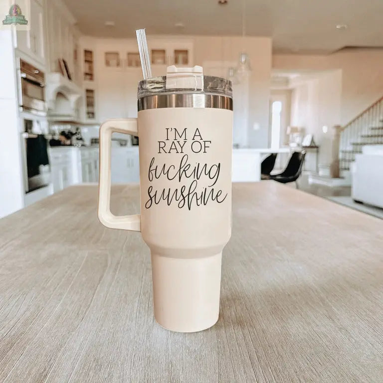The Ray of Sunshine 40oz cream-colored insulated mug with a straw sits on a wooden table in a bright kitchen, featuring black script and print that says, IM A RAY OF fucking SUNSHINE.