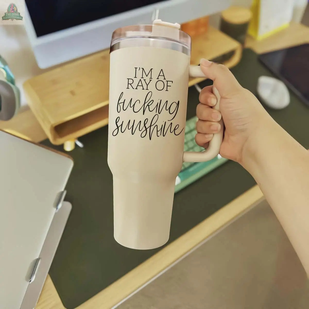 A hand holds the Ray of Sunshine 40oz beige insulated mug with "IM A RAY OF fucking sunshine" printed on it; a computer, laptop, and office supplies appear in the background.