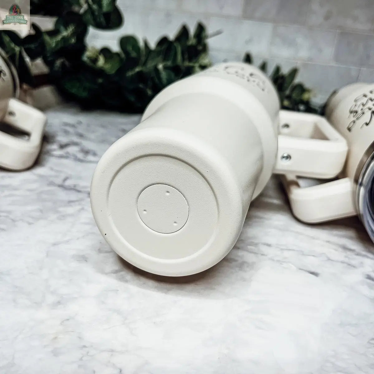 A close-up of the bottom of the Ray of Sunshine 40z white insulated mug tumbler on a marble countertop, with two similar travel mugs nearby and green foliage in the background.