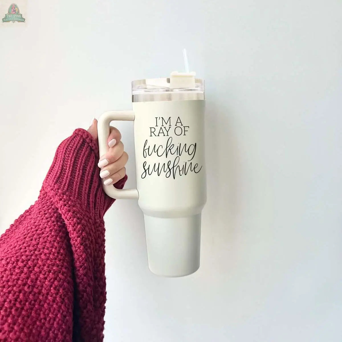 A person in a red knit sweater holds the Ray of Sunshine 40z—a large white insulated mug with "I'M A RAY OF f*cking sunshine" printed on it—against a plain, light-colored wall background.