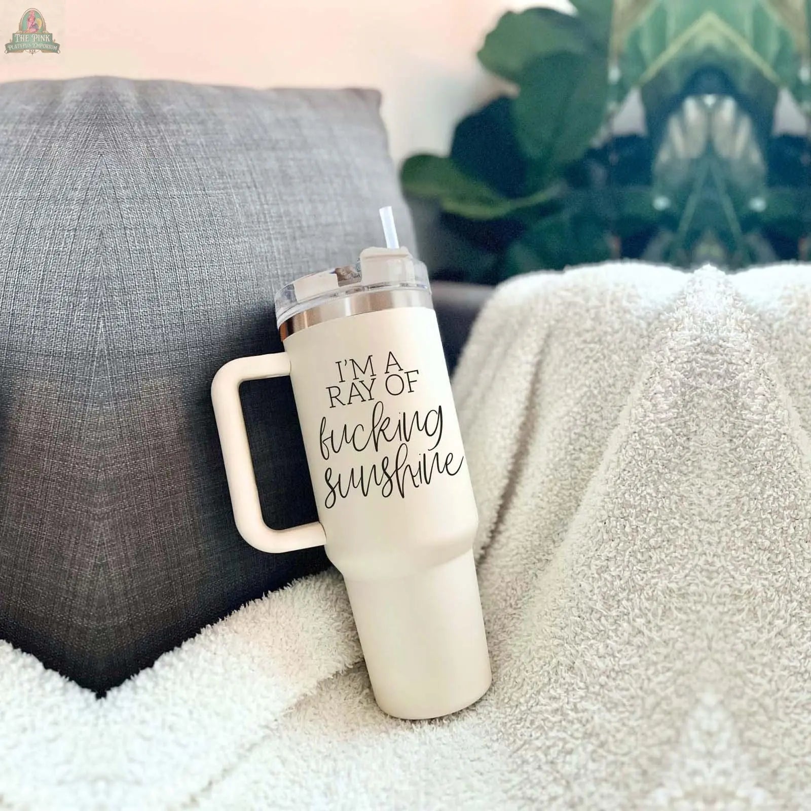 The Ray of Sunshine 40oz insulated mug, white with a handle and straw and "IM A RAY OF fucking sunshine" text, is shown resting on a white textured blanket beside a gray pillow and green leafy plants.