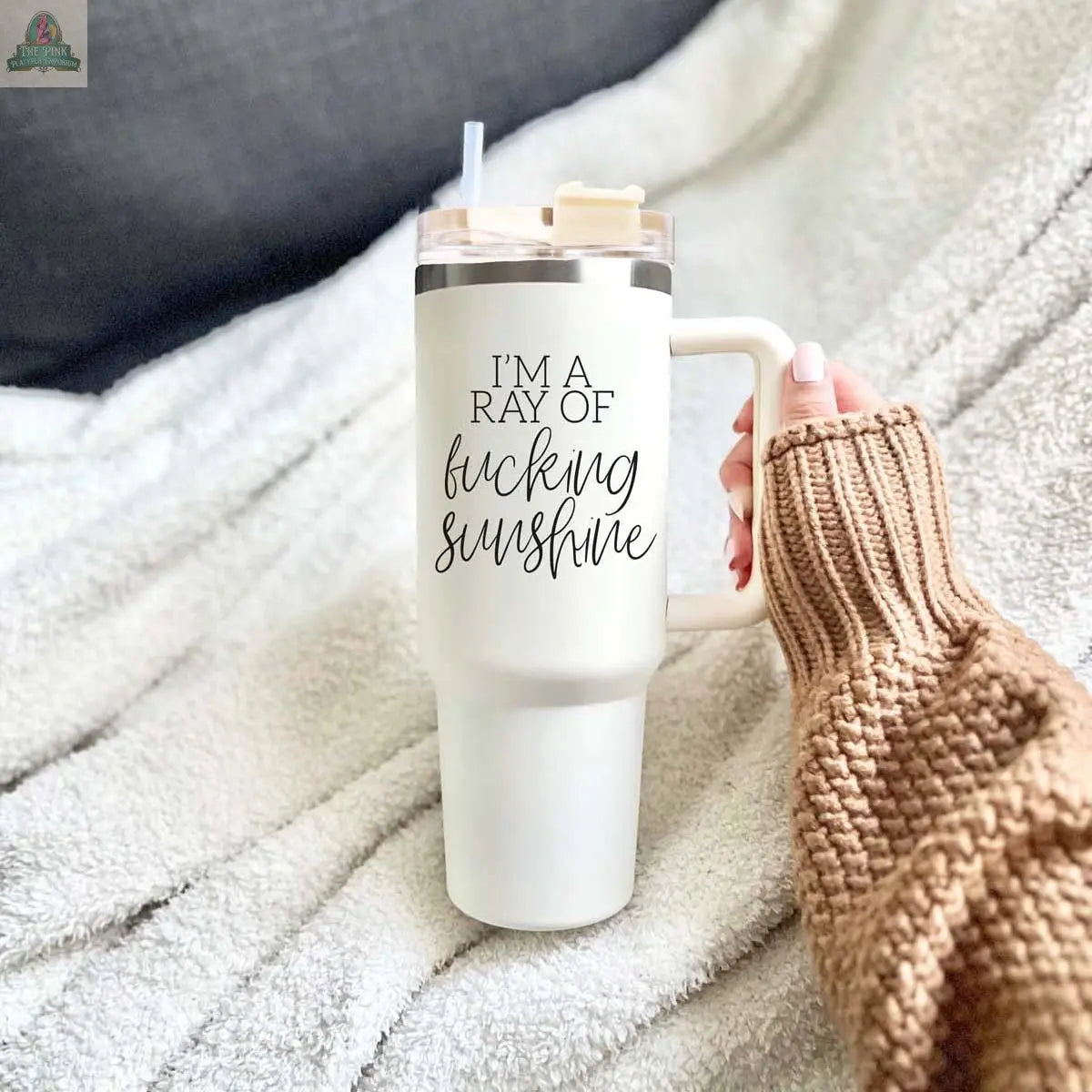 A hand in a brown knit sleeve holds the "Ray of Sunshine 40oz" white insulated mug with a straw, featuring the phrase "IM A RAY OF f*cking sunshine." A soft blanket appears in the background.