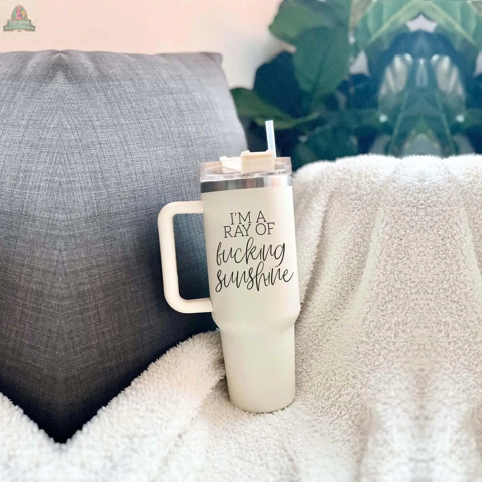 The Ray of Sunshine 40oz insulated mug, featuring IM A RAY OF f*cking sunshine, sits on a white blanket beside a gray pillow with green plants in the background.