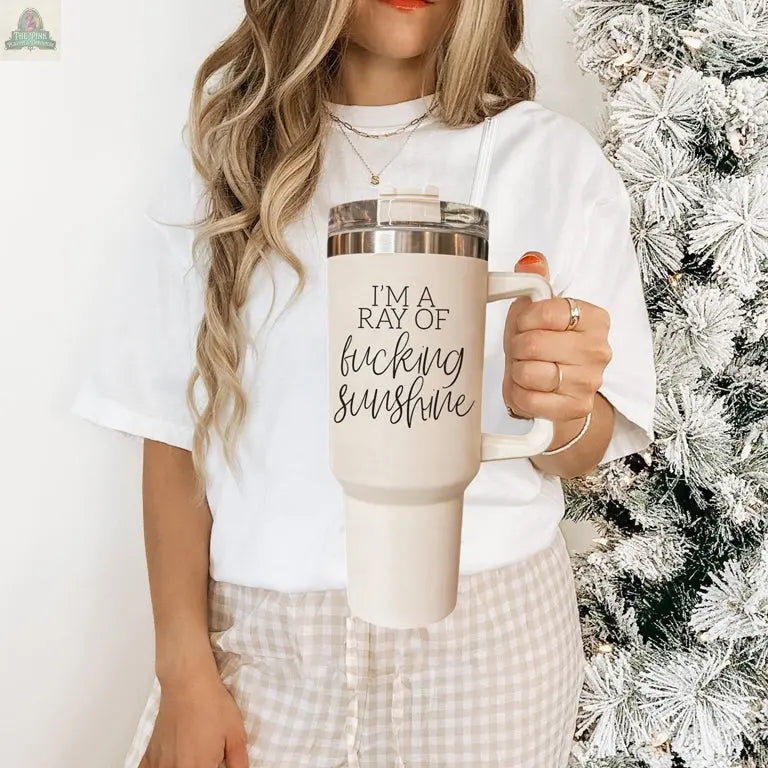 A woman with wavy light brown hair holds the Ray of Sunshine 40oz travel mug, featuring “I’M A RAY OF fucking sunshine.” She wears a white shirt and plaid pants, standing by a snowy artificial Christmas tree.