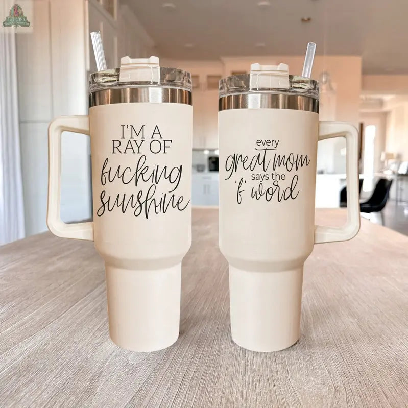 Two beige Ray of Sunshine 40oz insulated travel mugs with handles sit on a light wooden table; one reads “I’M A RAY OF fucking sunshine,” the other “every great mom says the f word.” Both come with clear lids and straws.