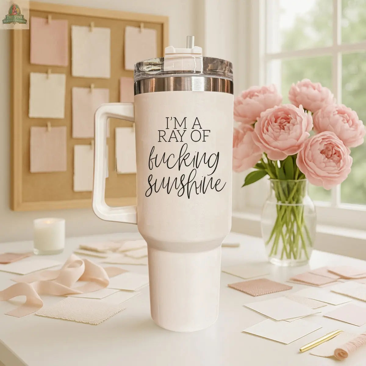 The Ray of Sunshine 40z insulated white mug featuring "IM A RAY OF fucking SUNSHINE" in stylish script sits on a desk surrounded by blush pink stationery, ribbon, and pink flowers near a sunlit window.