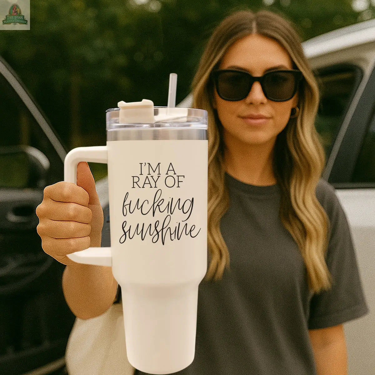 A woman in sunglasses and a gray shirt stands outdoors by an open car door, holding the Ray of Sunshine 40oz travel mug.