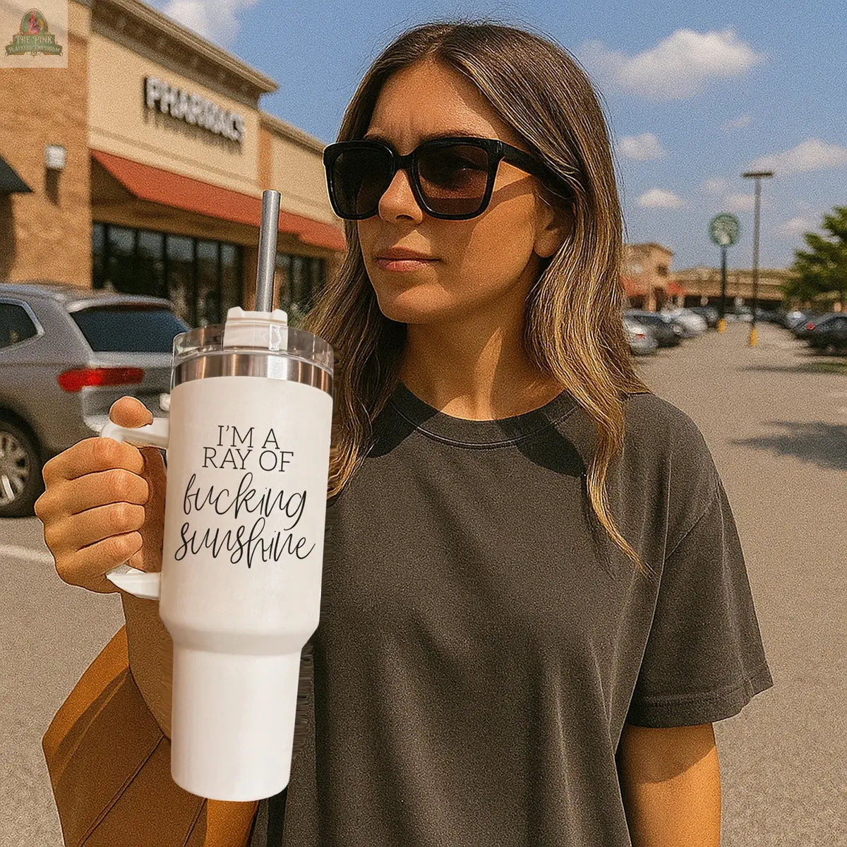 A woman in sunglasses stands in a parking lot holding the white Ray of Sunshine 40oz mug featuring "I’m a ray of fucking sunshine." She wears a dark t-shirt, with cars and shops visible behind her.