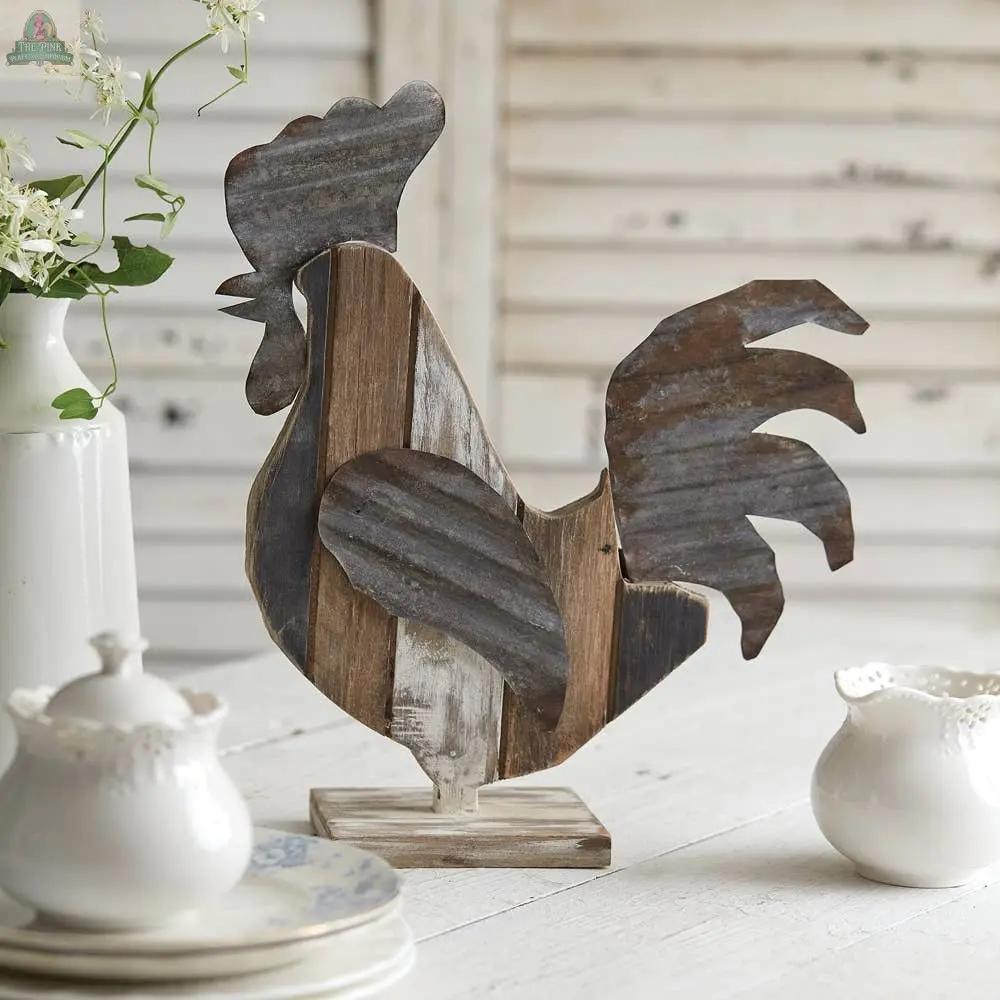 The Reclaimed Wood Rooster sits on a wooden table, surrounded by white ceramic dishes and a flower vase, adding farmhouse charm to a white shiplap setting.