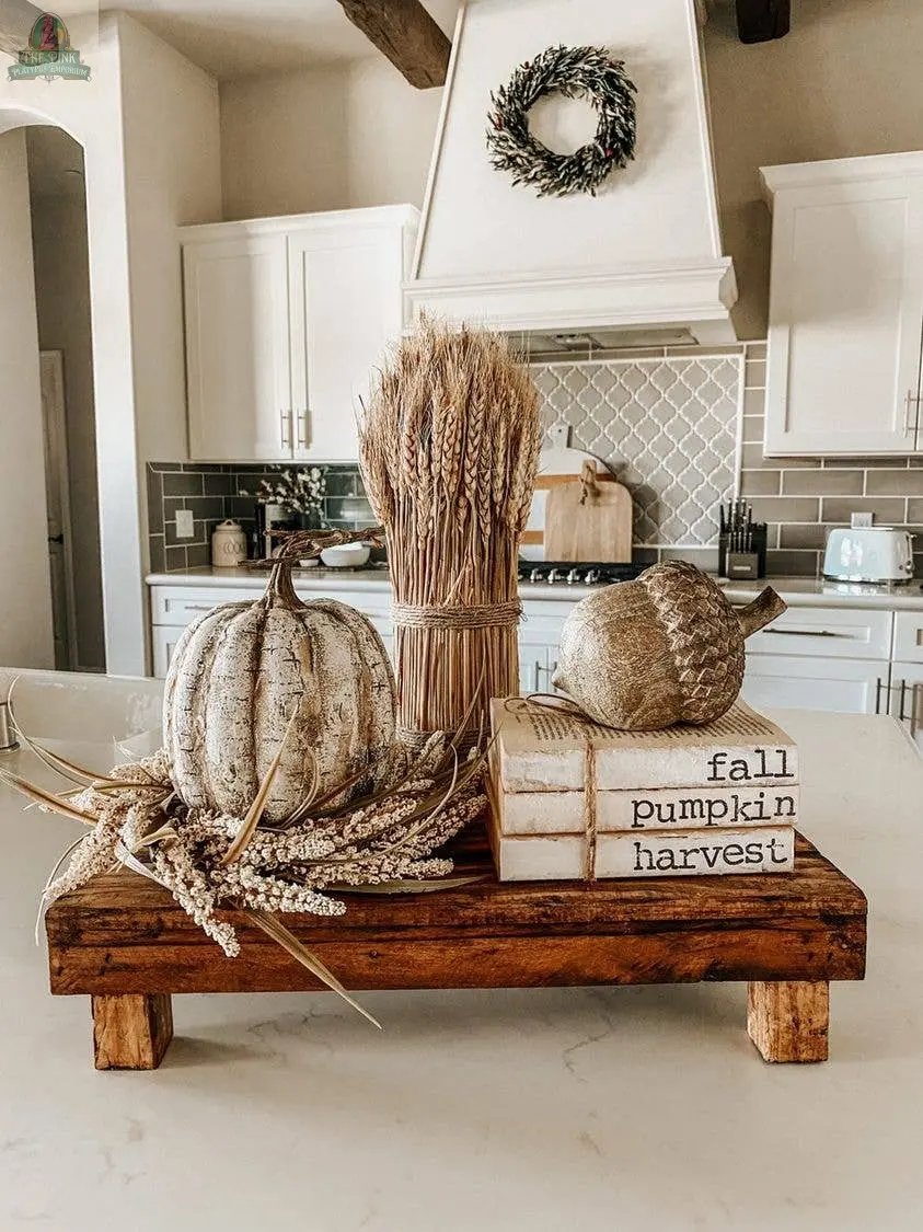 A rustic fall centerpiece on a handmade rectangular wood stool features white pumpkins, dried corn, wheat stalks, and books labeled fall, pumpkin, and harvest, set on a bright kitchen island with a wreath above the stove.