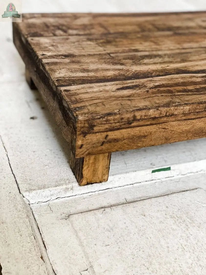 A close-up of the Rectangular Wood Stool, Natural, Handmade, featuring short block legs and a weathered finish with visible grain, shown on a textured white surface—ideal for adding rustic charm to your home decor.