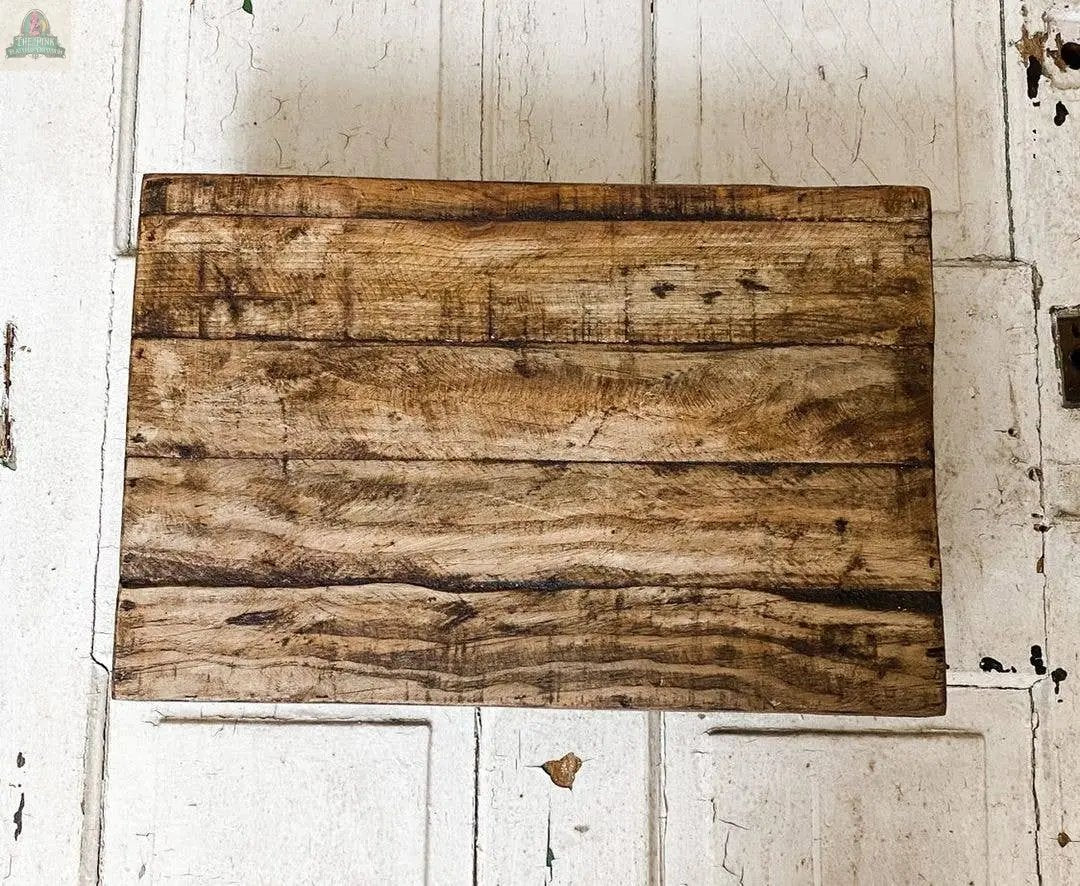 A handmade rectangular wood stool in a natural finish with a rustic, weathered texture rests on a white, distressed wooden surface.