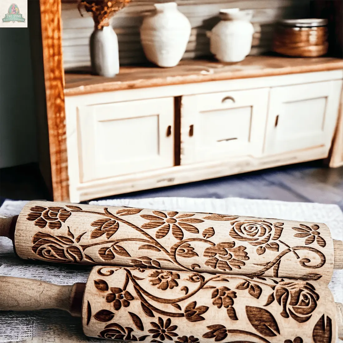 Two Rolling Pins - Box Style with intricate floral carvings rest on a table, making unique baking gifts. In the background, a rustic wooden cabinet and pottery add extra charm.