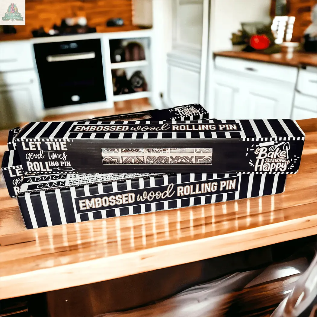 A Rolling Pins - Box Style embossed wood rolling pin, packaged in a black and white striped box with decorative text, sits on a kitchen counter—perfect as a baking gift. A modern kitchen is seen in the background.