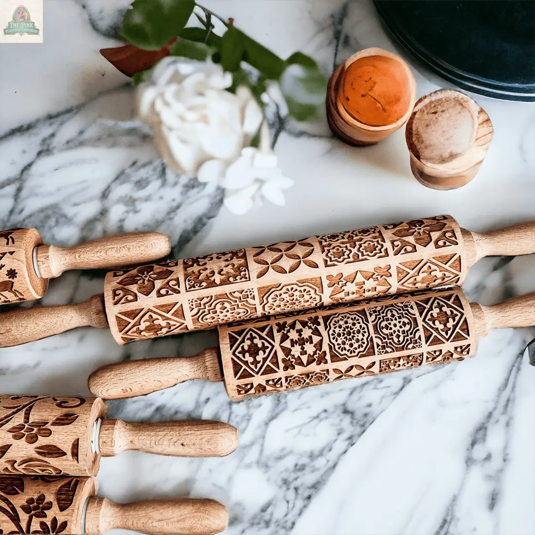 Two "Rolling Pins - Box Style" with intricate carvings rest on marble near a white flower, greenery, a dark green dish, and wooden spice containers—ideal baking gifts for those who appreciate unique kitchen tools.
