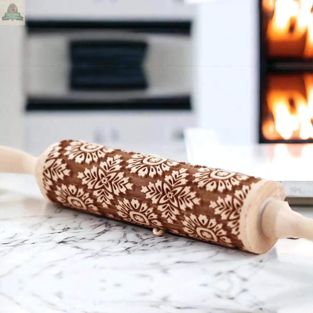 A Rolling Pins - Box Style rests on a marble countertop in a bright kitchen, displaying intricate floral and snowflake designs—ideal for baking gifts. A glowing fireplace adds warmth in the background.