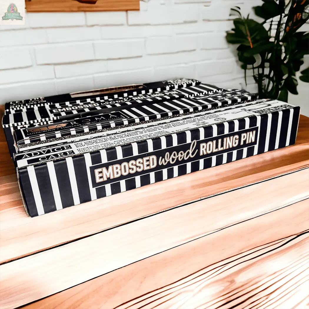 A striped black and white box labeled "Rolling Pins - Box Style" rests on a wooden surface, ideal as a unique baking gift, with a white brick wall and a green plant visible in the background.