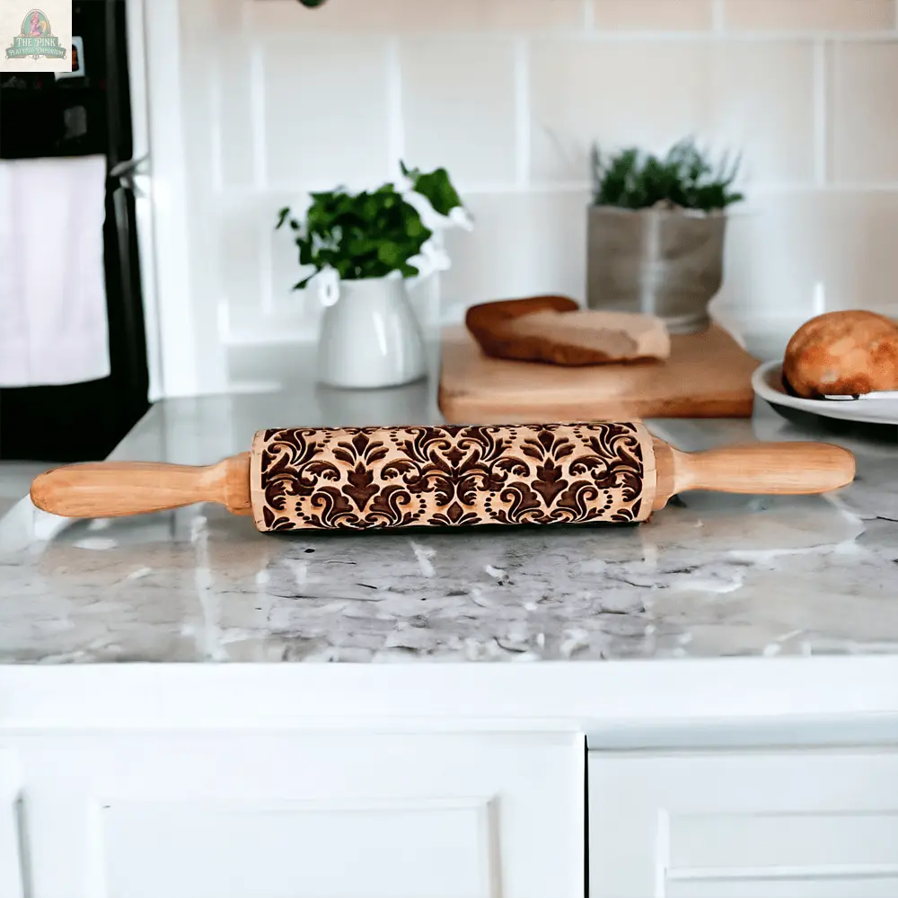 The Rolling Pins - Box Style, featuring an intricate floral design, sits on a marble kitchen counter, ideal for baking gifts. Houseplants, cookie cutters, bread on a cutting board, and a white tiled wall appear in the background.