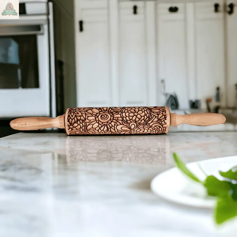 The Rolling Pins - Box Style features an intricate floral pattern and rests on a marble countertop with green leaves—a perfect unique baking gift. A blurred white plate in the foreground adds a touch of elegance to the scene.